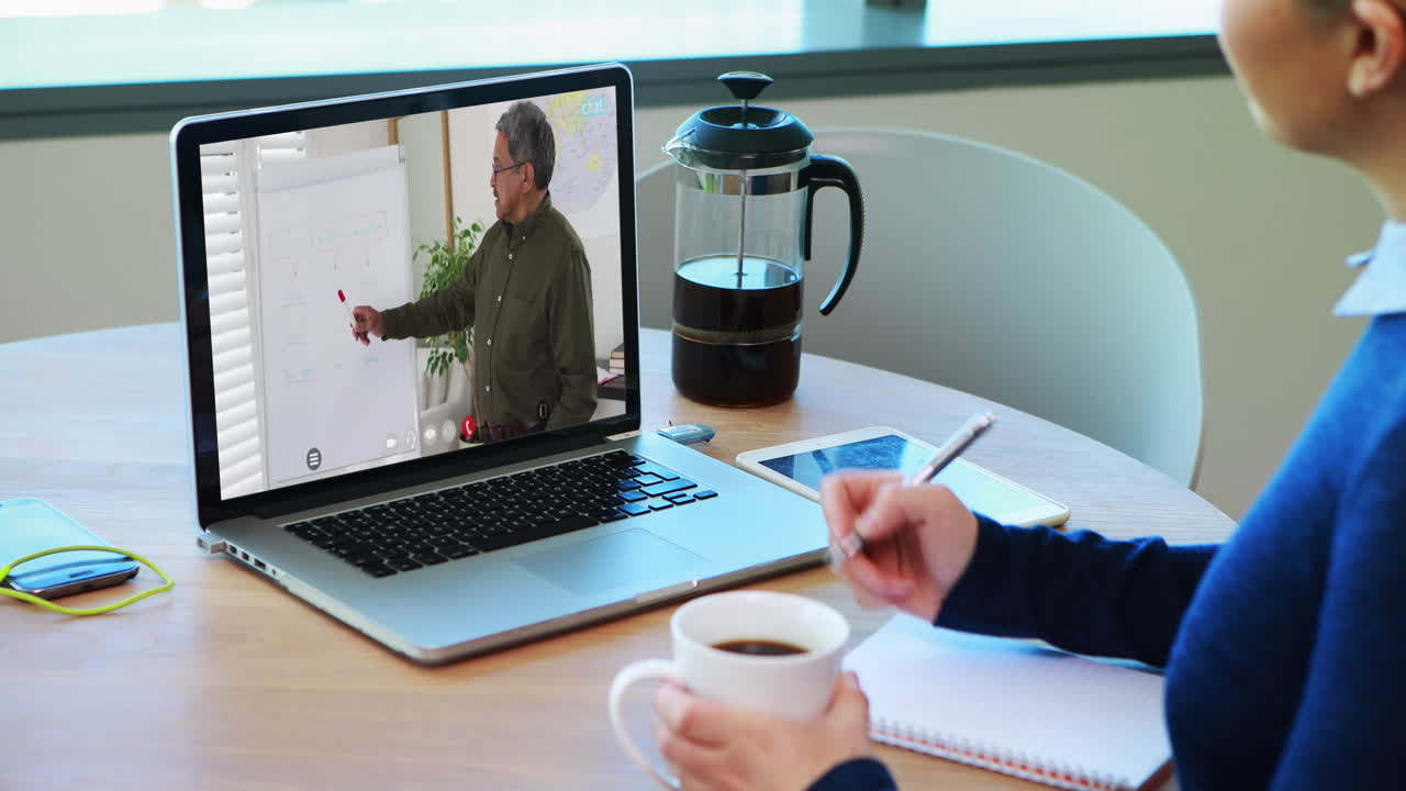 Caucasian female student using laptop on video call with male teacher, making notes
