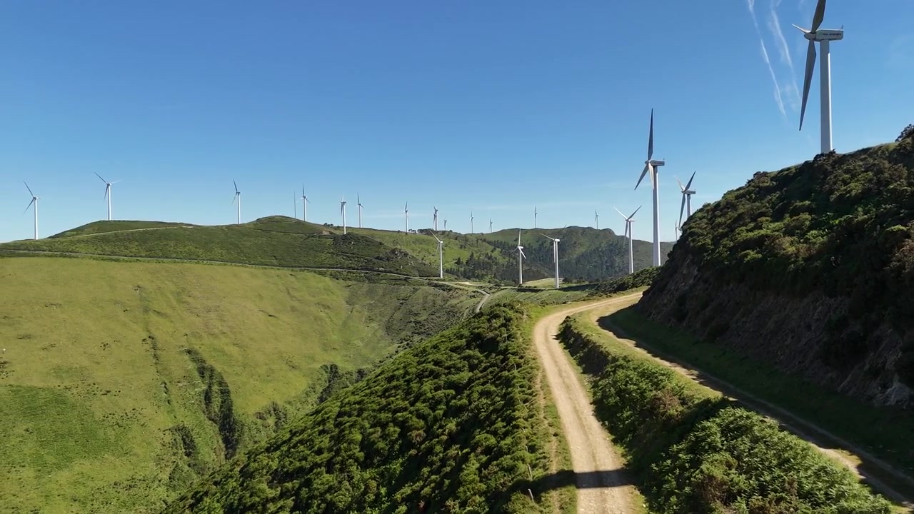 Vista drone lungo una strada sterrata che costeggia turbine su un verde pendio di montagna.