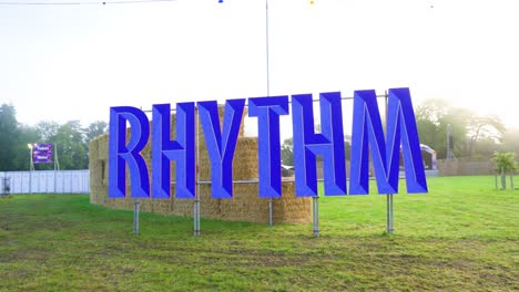 huge rhythm text sign of blue colour realistic 3d with inward lines placed hanging on metal fence in the nature on grassy field sunrise clear sky clouds plenty bales on top of each other festival area