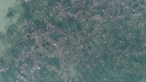 High-altitude-top-down-descending-drone-shot-of-polluted-water-filled-with-trash-floating-over-dead-coral-reef-in-the-turqouise-tropical-water-of-Bali-Indonesia