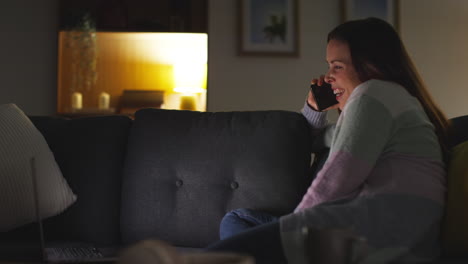 Smiling-Woman-Sitting-On-Sofa-At-Home-At-Night-Talking-On-Mobile-Phone-And-Watching-Movie-Or-Show-On-Laptop-3