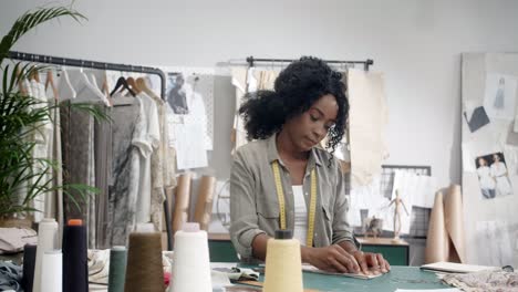 joven diseñadora de ropa sentada en su estudio en la mesa y haciendo algunas medidas en la tela