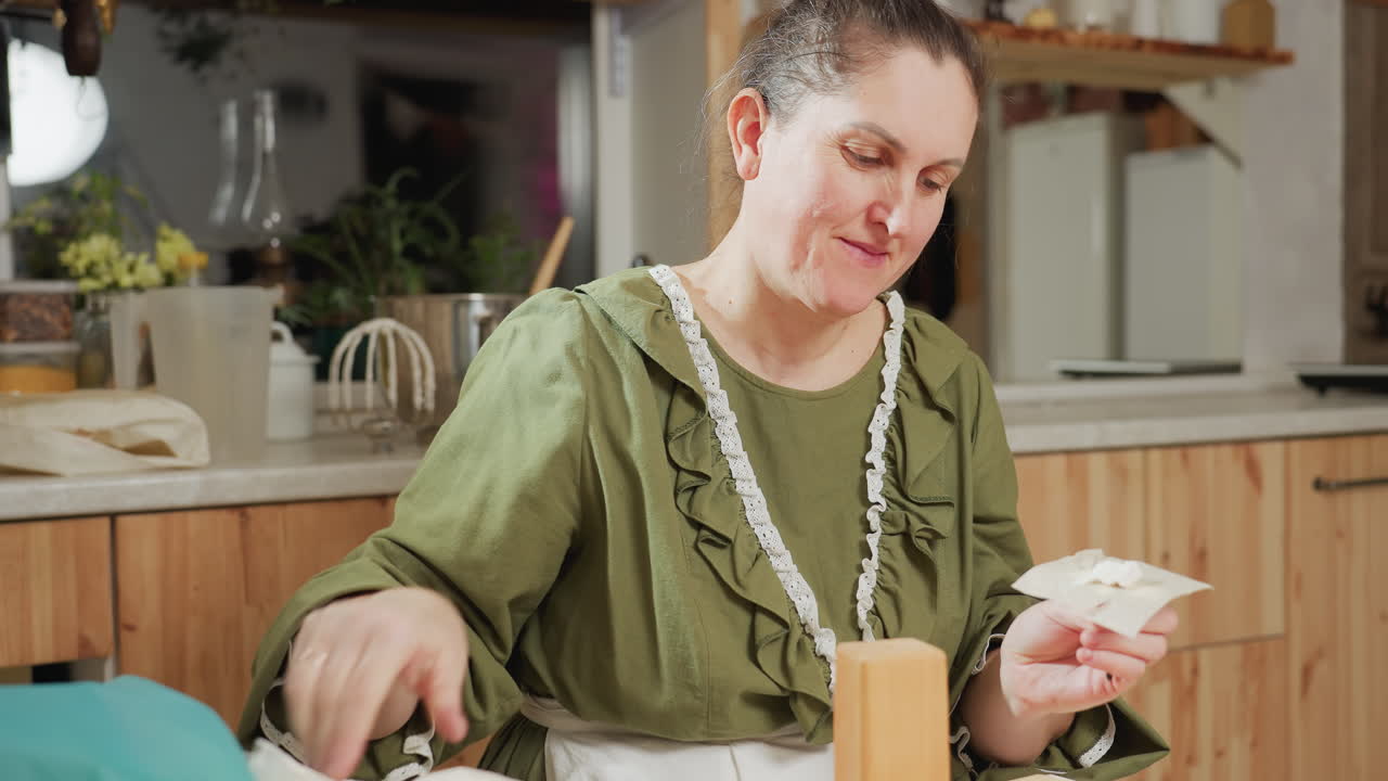 Woman in green outfit places white icing onto cut out paper on fooddart, then adds dessert on top while baking in cozy kitchen setting, enjoying moment and smiling warmly as she licks icing from hand