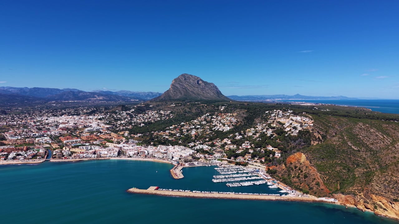 Città costiera di Javea sulla Costa Blanca spagnola con porto turistico, barche ormeggiate, mare Mediterraneo turchese e il monte Montgó che si erge dietro ville bianche sulla collina.