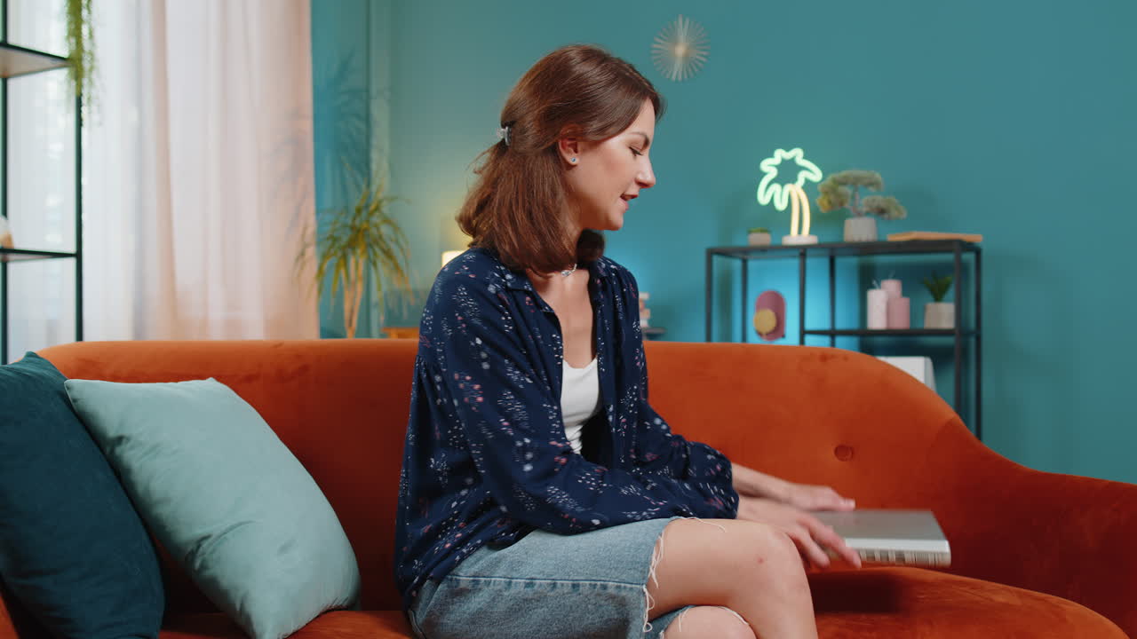 행복한 백인 젊은 여성이 소파에 앉아 일을 마친 후 노트북을 닫고 집에서 휴식을 취하고 있습니다.