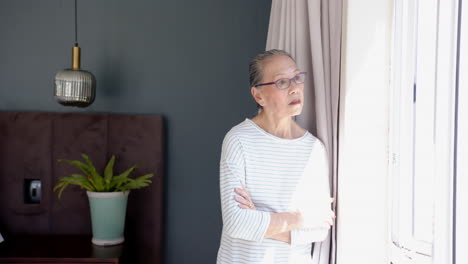 looking out window, elderly woman in striped shirt standing with arms crossed