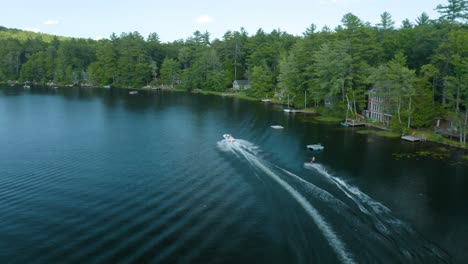 Outdoor-shot-of-a-person-water-skiing-at-a-blue-lake-with-splashes-of-water-or-waves,-many-lush,-green-trees-in-the-background,-a-few-residences,-and-overcast-sky-can-be-seen-in-the-distance