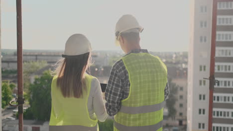 Vista-Posterior,-Joven-Capataz-E-Ingeniero-Revisando-El-Plano-Y-Hablando-Sobre-El-Proyecto-De-Construcción-Con-Compromiso-Con-El-éxito-En-El-Sitio-De-Construcción