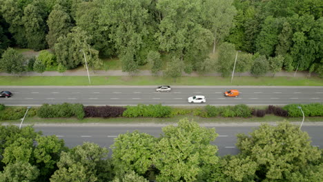 aerial top down - flying a drone parallel to the road lane - multi-lane roadway with moving vehicles - wide street in the city between trees and greenery