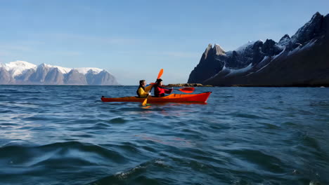 two people kayaking in a red boat on open water with snowy mountains in the background