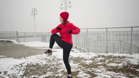 back view of woman performing energetic winter workout outdoors on snowy ground near railing overlooking water, wearing red hoodie and black gloves, with foggy atmosphere and serene background