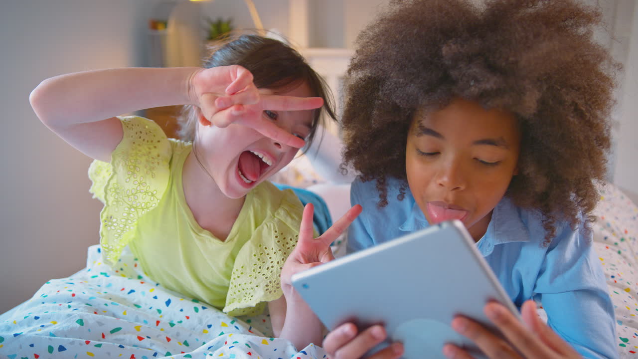Girl And Boy In Bedroom Lying On Bed Using Digital Tablet And Pulling Funny Faces Together