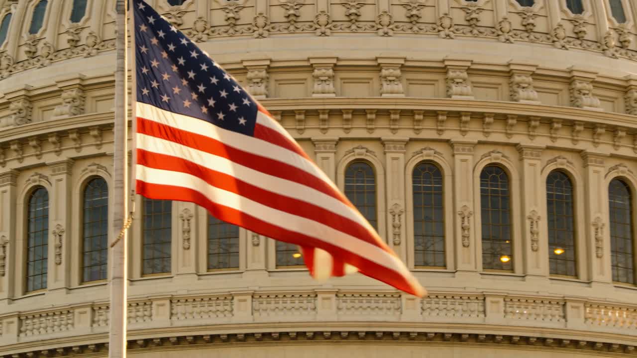 Bandiera americana che sventola, le raffiche la spingono a srotolarsi e ondeggiare sull'asta davanti a una facciata a cupola con strisce illuminate.