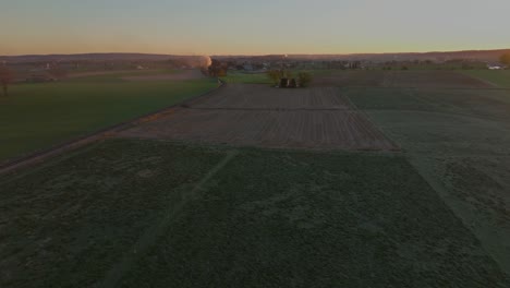 an aerial moving view of a sunrise over farmlands, barns, and silos as a steam train approaches in the distance on an autumn morning