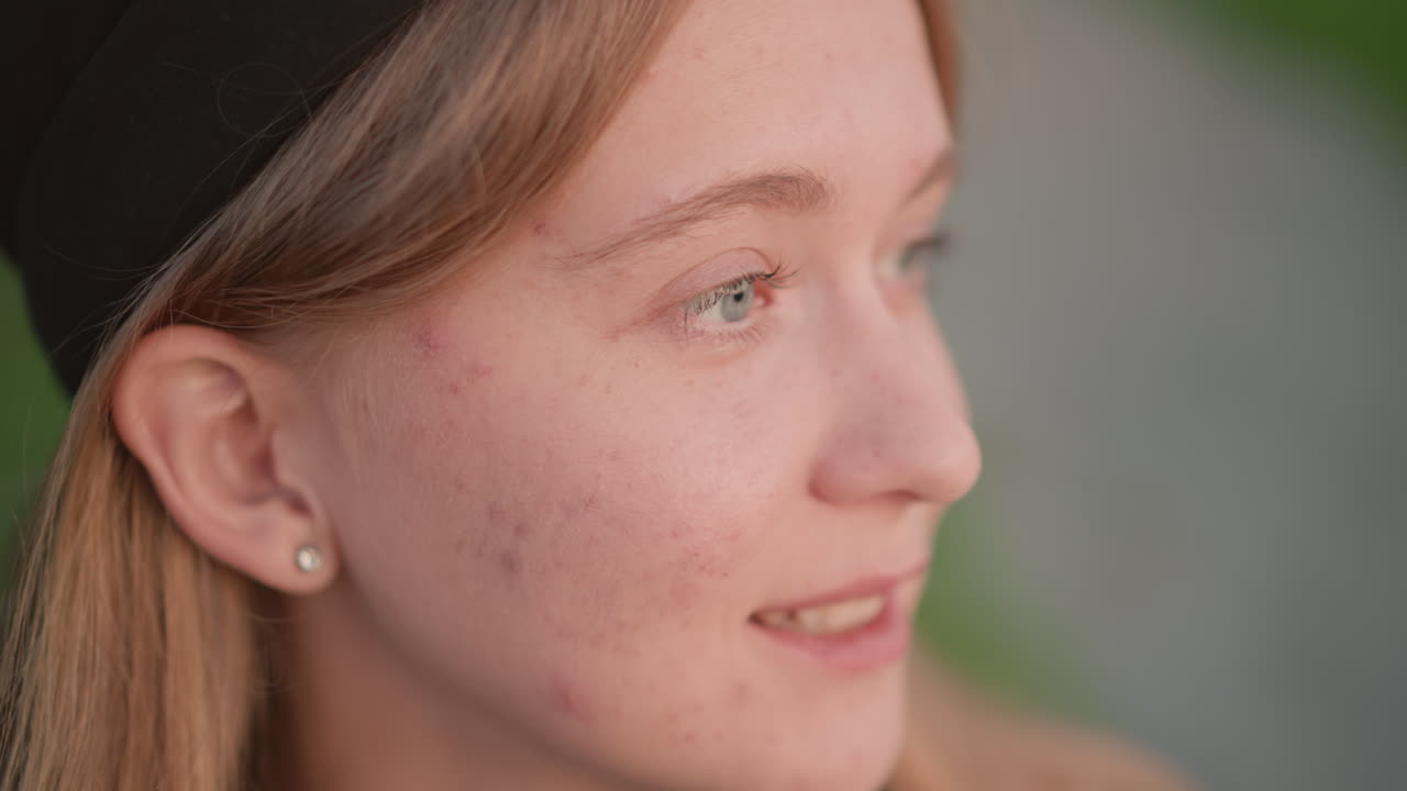 sorriso spontaneo di una donna bianca con le cuffie in un parco, sguardo speranzoso verso il sentiero e l'orizzonte verdeggiante, postura rilassata che suggerisce un'artista di strada o una ballerina che pianifica la prossima mossa, tenue bagliore serale, orecchino che cattura la luce