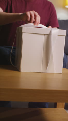 vertical video of man opening present in gift wrapped box decorated with ribbon on table in lounge