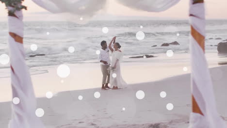 animation of white spots over african american bride and groom dancing on beach at wedding