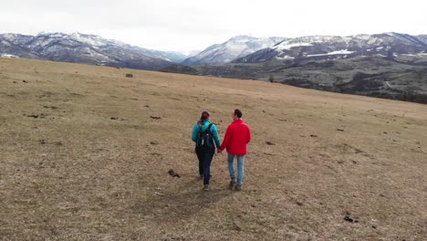 drone follows a couple walking hand in hand across open countryside in armenia with mountains in the background as they walk through a grassy field on a bright day