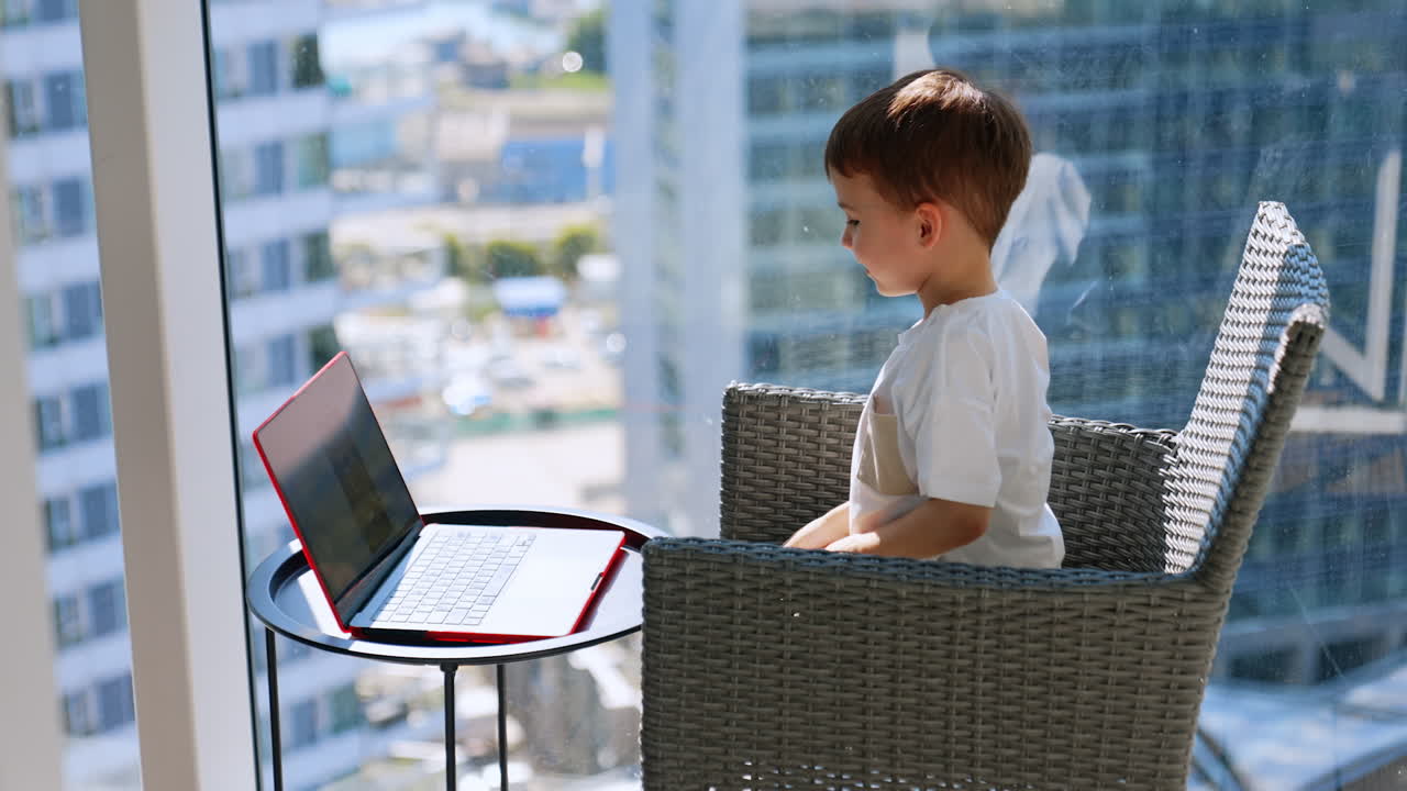 Lovely child sits in the chair near panoramic window on sunny day. Happy kid uses laptop standing on the coffee table in front of him