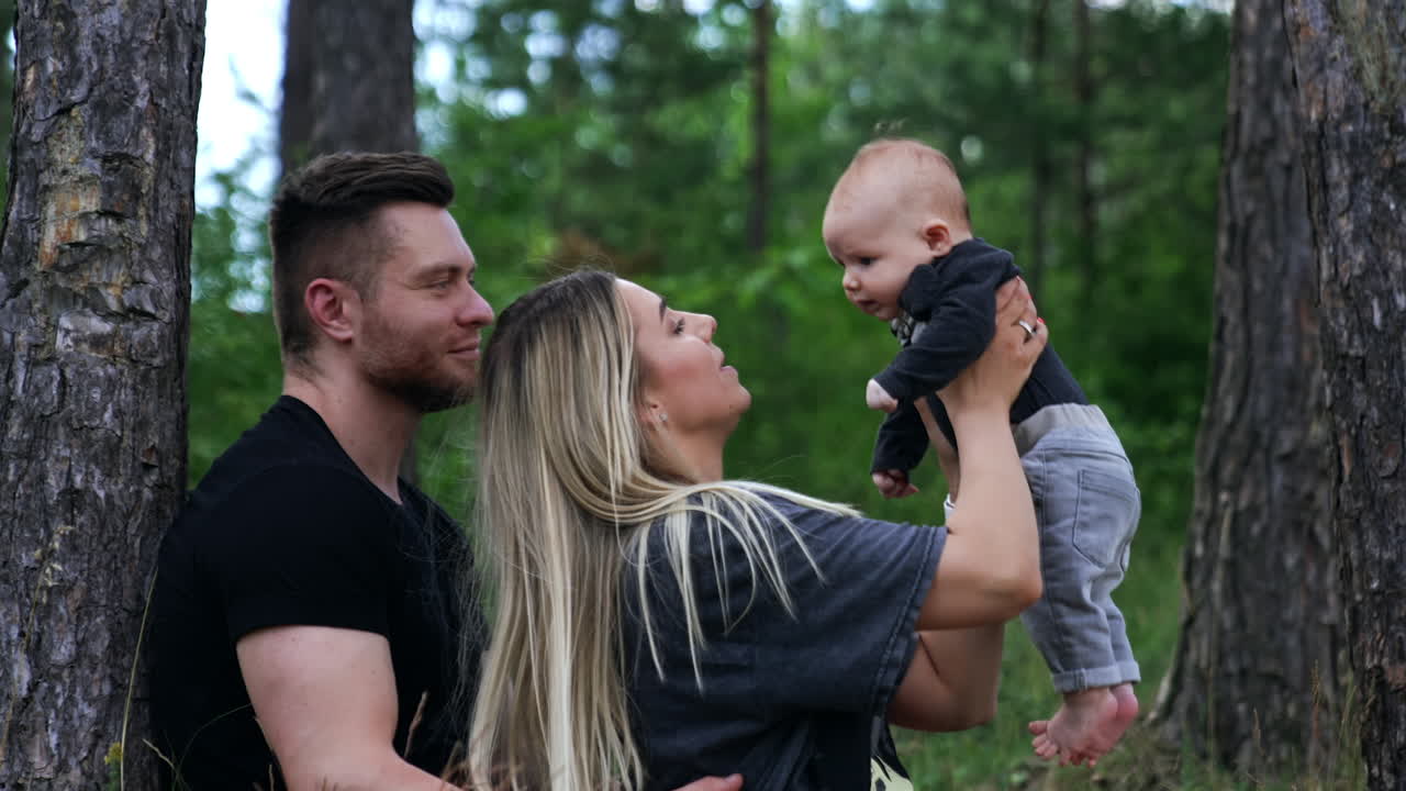 Family of three spending time in the nature. Woman holds her child above the head and kid smiles adorably.