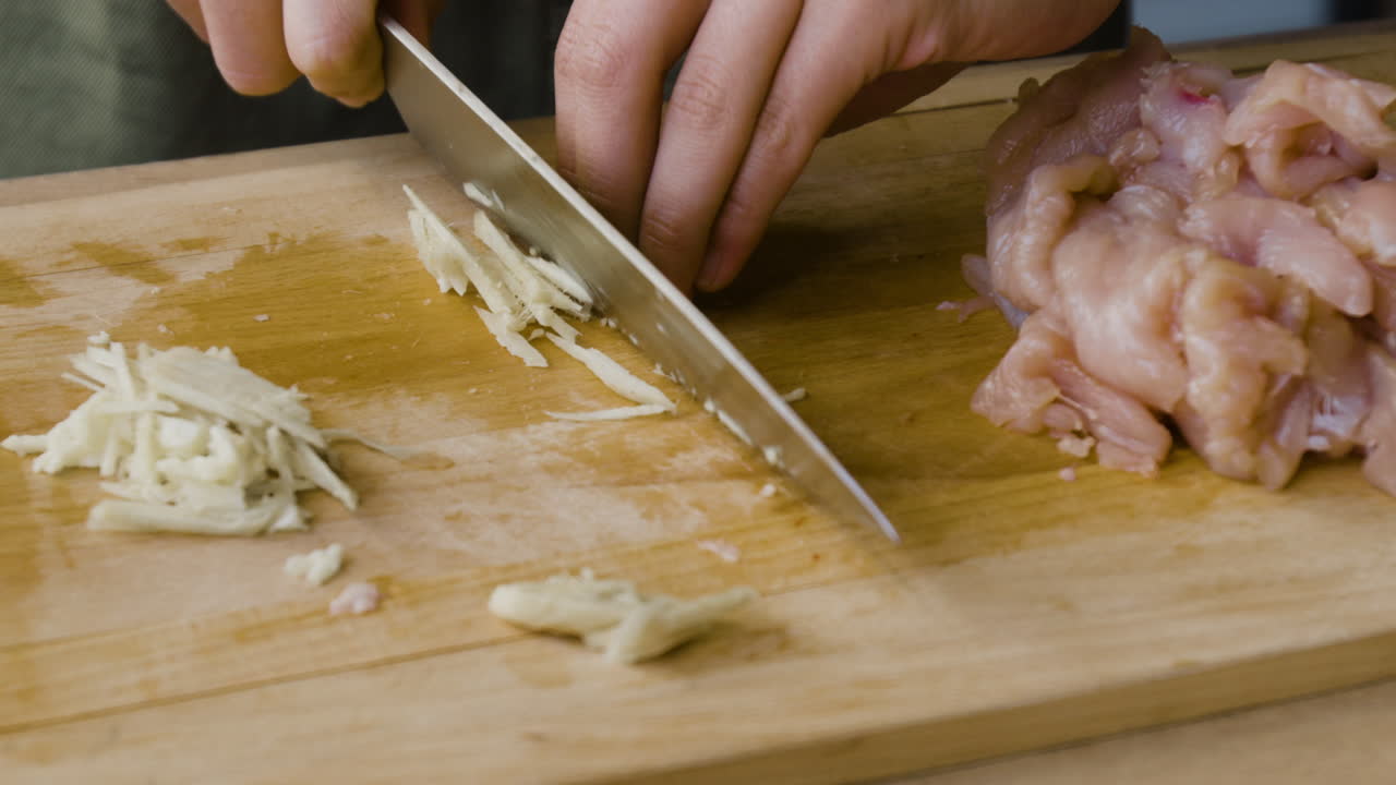A close-up of hands chopping ingredients on a cutting board next to raw chicken