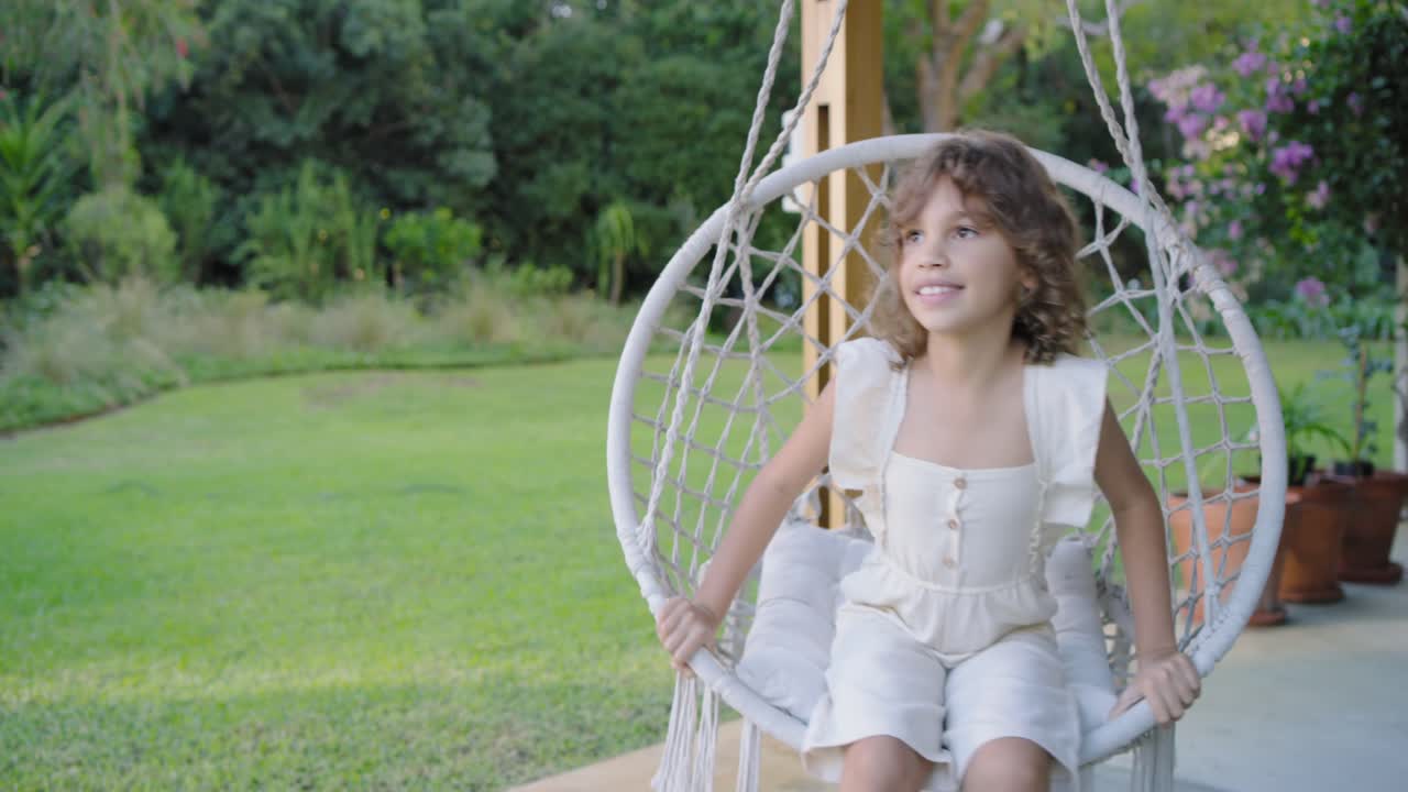 Bambino afroamericano che arriva e si sistema su una sedia in macramè in veranda, in tutina, spingendo le corde per dondolarsi.