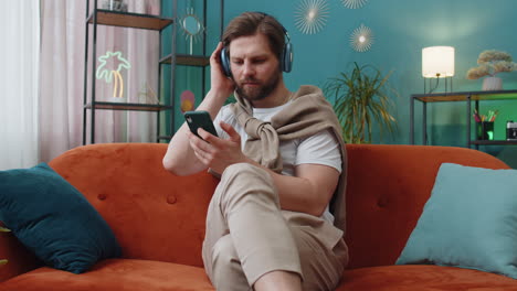 happy caucasian man in wireless headphones relaxing sitting on sofa at home listening favorite music