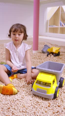 little girl playing with yellow toy dump truck in indoor playground filled with small bright pellets