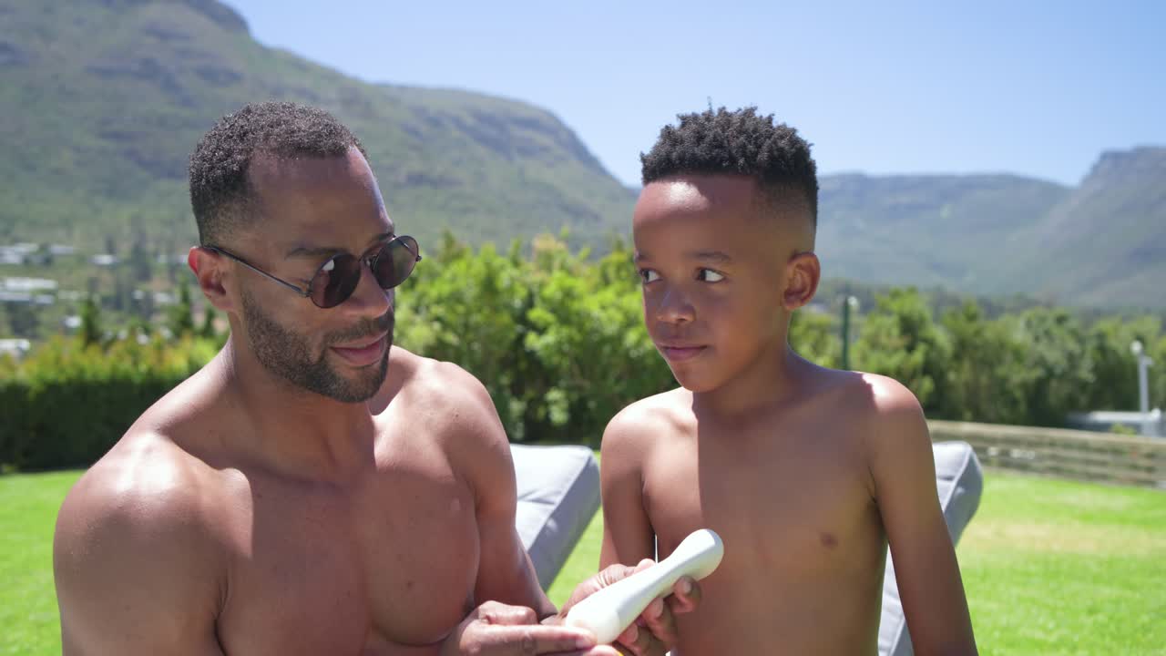 Father and son sitting on lounge chair, pointing at nose and applying sunscreen, protecting skin