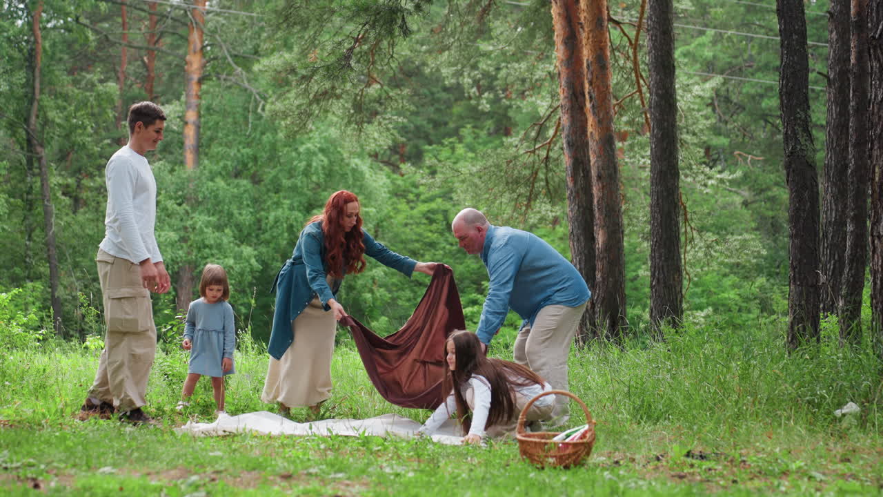 아이들이 우유 색깔 천을 펼치고 부모는 갈색 천을 펼치는 동안, 숲에서 즐거운 가족이 피크닉 장소를 준비하는 모습을 멀리서 촬영했어요. 어린아이가 호기심 가득한 눈으로 지켜보고 있으며, 주변은 푸른 녹음으로 가득해요.