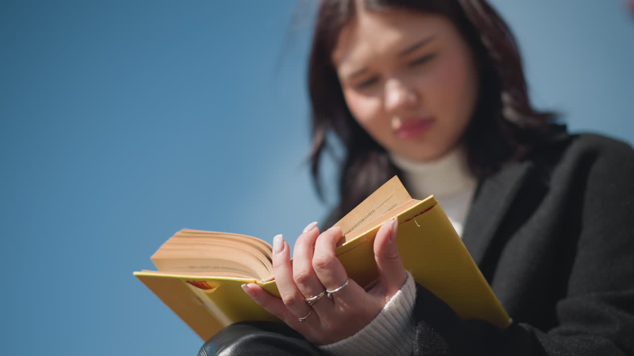 Close-up of student with well-polished nails adorned with rings, thoughtfully reading and observing pages of yellow cover book, contemplating outdoors in bright sunlight