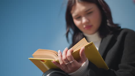 close-up of student with well-polished nails adorned with rings, thoughtfully reading and observing pages of yellow cover book, contemplating outdoors in bright sunlight