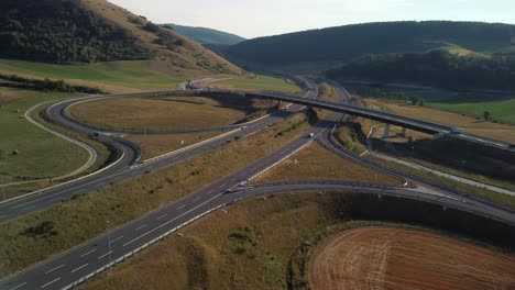 Vista-Aérea-De-La-Red-De-Carreteras-Con-Cruces-De-Entrada-Y-Salida-De-Autopista-En-Una-Zona-Montañosa-Al-Atardecer