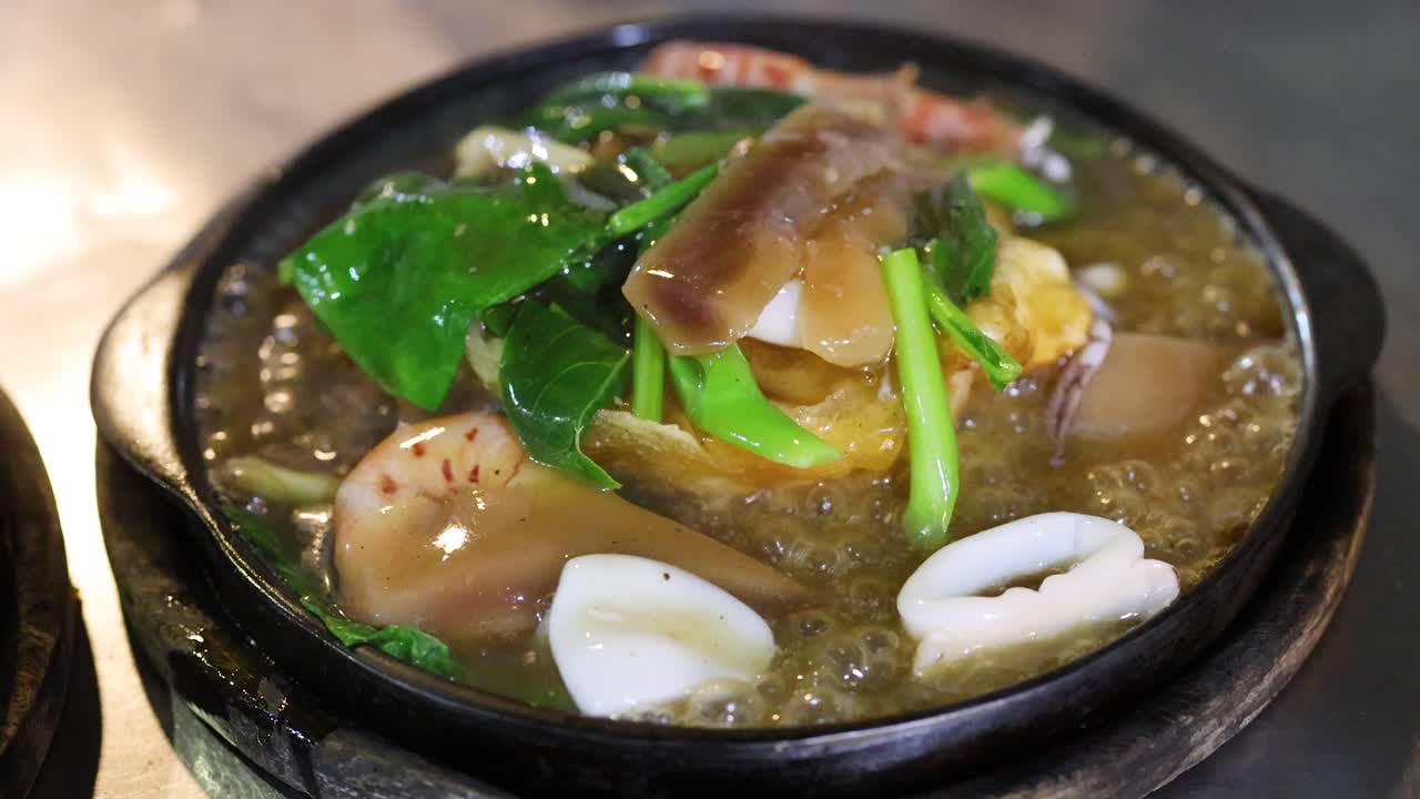 Seafood noodles simmer and bubble on a hot plate under warm lighting, close-up, minimal movement