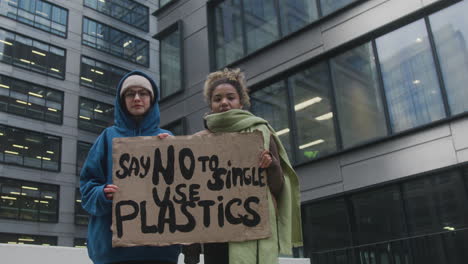 Zwei-Junge-Klimaaktivistinnen-Halten-Ein-Banner-Hoch-Und-Protestieren-Gegen-Einwegkunststoffe,-Während-Sie-In-Die-Kamera-2-Schauen