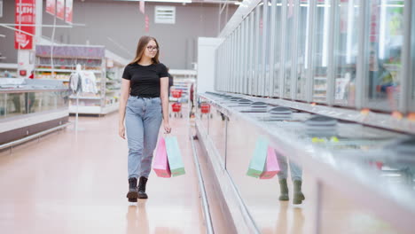 young lady walking through mall holding shopping bags, looking at glass display with reflection seen on sleek cabinets, a signpost hanging in the background and another person passing in the distant