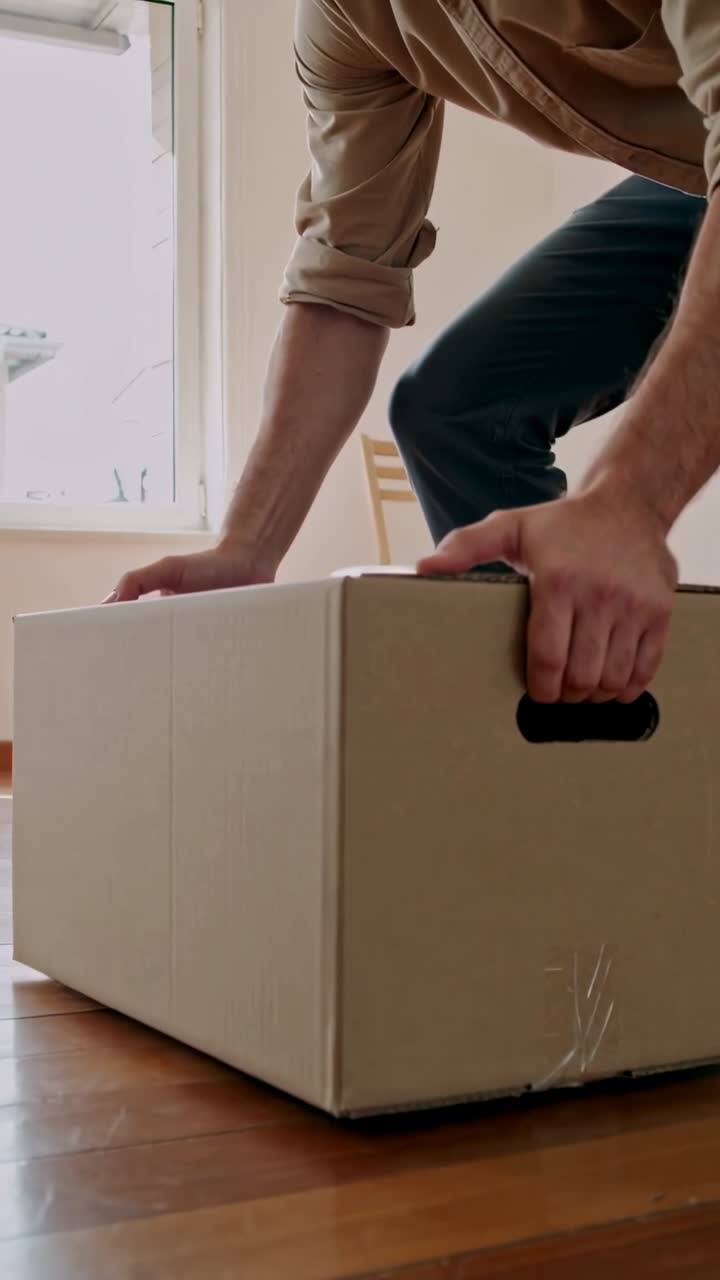 Vertical video: Walking in carrying moving box, man in jeans, boots lowering box on floor to unpack