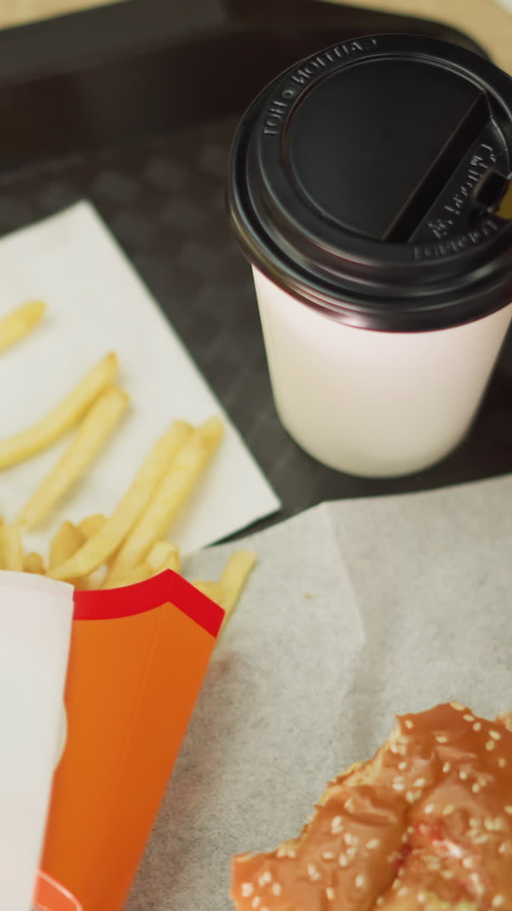 un primer plano de patatas fritas, una hamburguesa parcialmente comida, y una taza de café en una bandeja negra una mesa de madera marrón, la mano de una mujer en un vestido rosa alcanza las patatas frites, toma un bocado, y beba su café