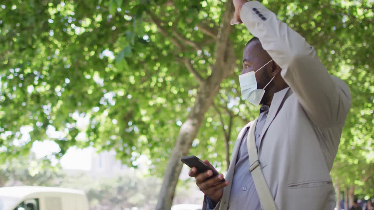 African american man in city wearing face, using smartphone and waving