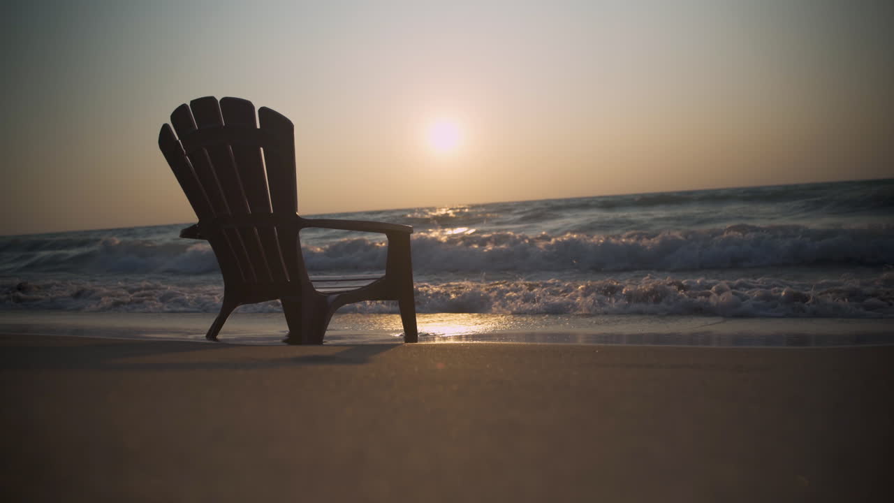 An empty Muskoka chair sits on a beach as the sun sets in the background