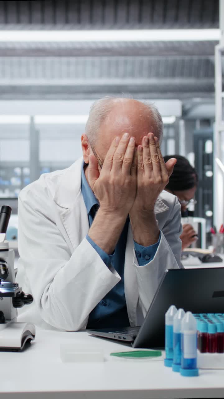 Video verticale: ricercatore anziano nel campo della nanotecnologia di laboratorio si sente sopraffatto mentre lavora al laptop