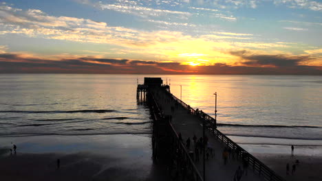 drone shot rising over newport beach pier with many people walking in silhouette at sunset over the california coast aerial