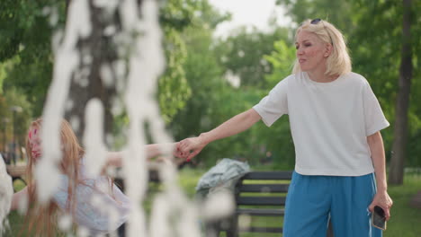 exciting moment between mother and daughter in park as girl grabs mothers hand guiding it into fountain water while mother smiles holding phone, green trees background, summer bonding