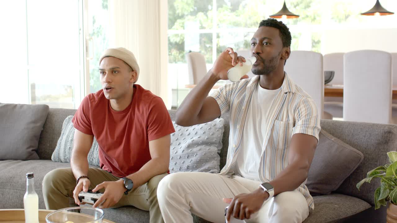 Diverse male friends settling on sofa at home, grabbing gamepads, sipping drinks and competing