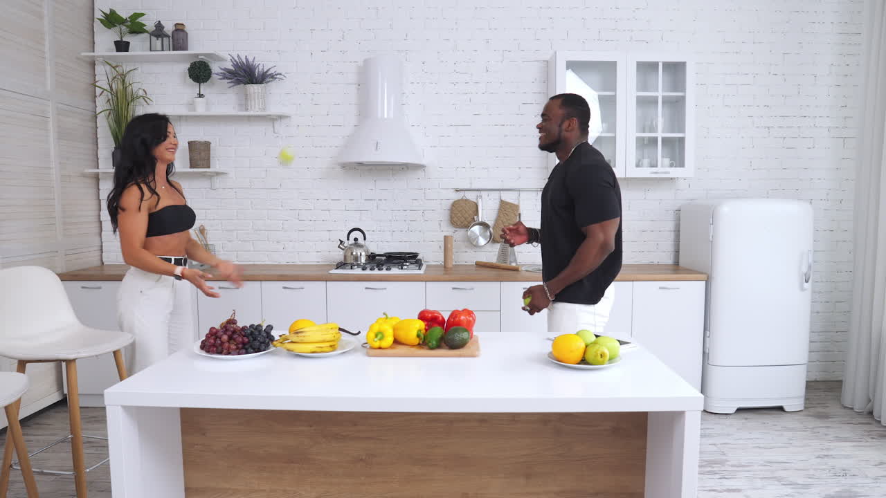 Happy couple having fun in their kitchen. Man and woman juggle with fruit and laughing. Light modern kitchen at the backdrop.