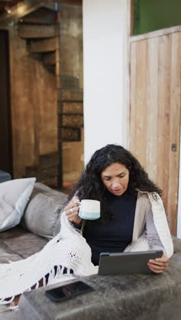 vertical video of biracial woman sitting on sofa and using tablet at home, slow motion