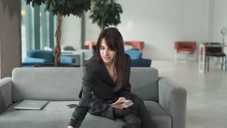 una mujer joven con un traje elegante se sienta en el vestíbulo de un centro de negocios y con alegría cuenta dinero - billetes de dólar