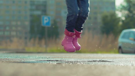 funny girl in pink rubber boots has fun jumping in puddle