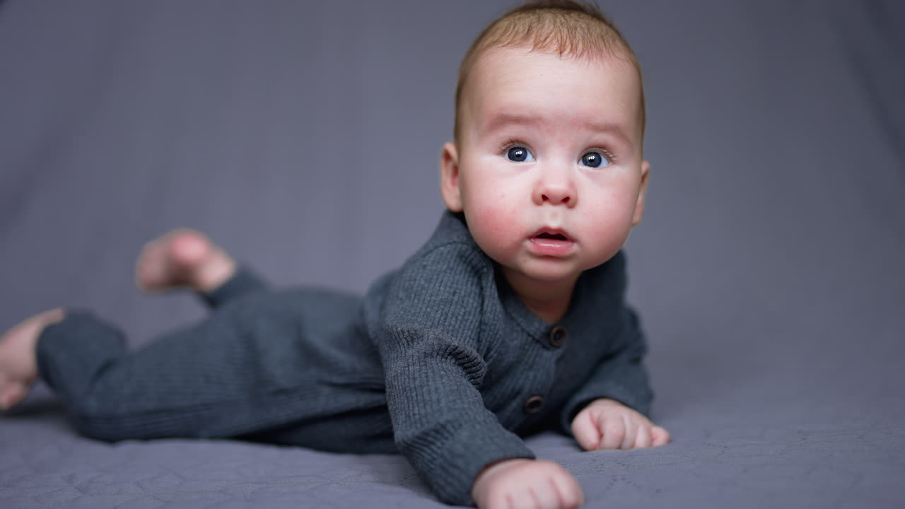 Healthy lovely kid in grey clothes lies on his belly. Adorable child looking sideways with interest. Grey blurred backdrop.