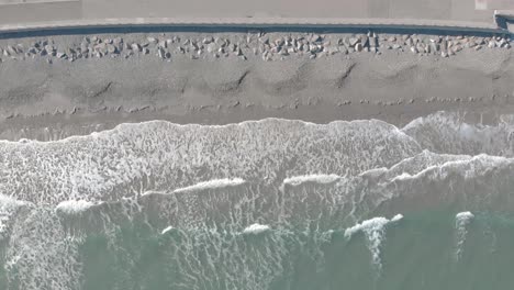 static birds-eye aerial of waves crashing softly on the sandy shoreline next to a cement seawall